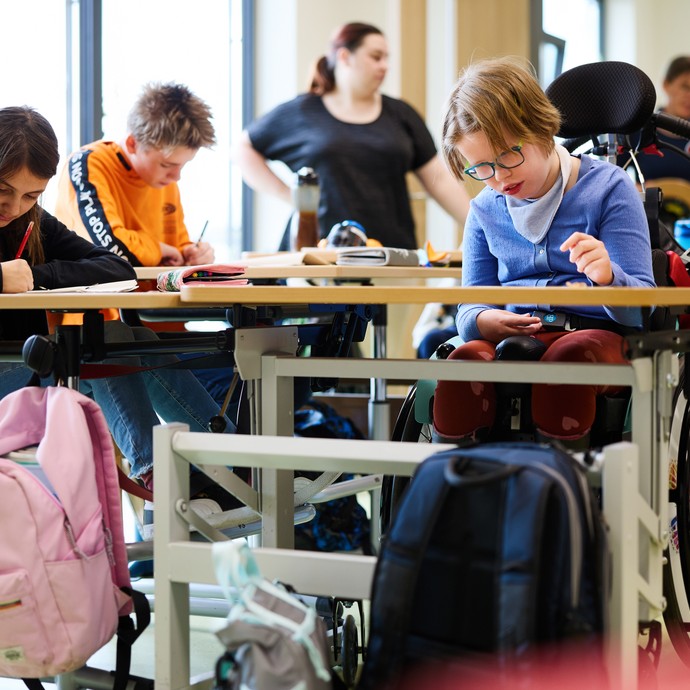 Kinder sitzen in einem Klassenraum an ihren Tischen und arbeiten. Ein Mädchen sitzt im Rollstuhl. (vergrößerte Bildansicht wird geöffnet)