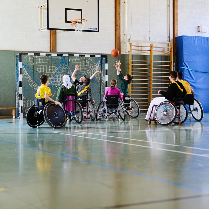 Acht Kinder in Rollstühlen stehen in einer Sporthalle unter einem Basketballkorb. Ein Mädchen versucht den Ball in den Korb zu werfen. (vergrößerte Bildansicht wird geöffnet)