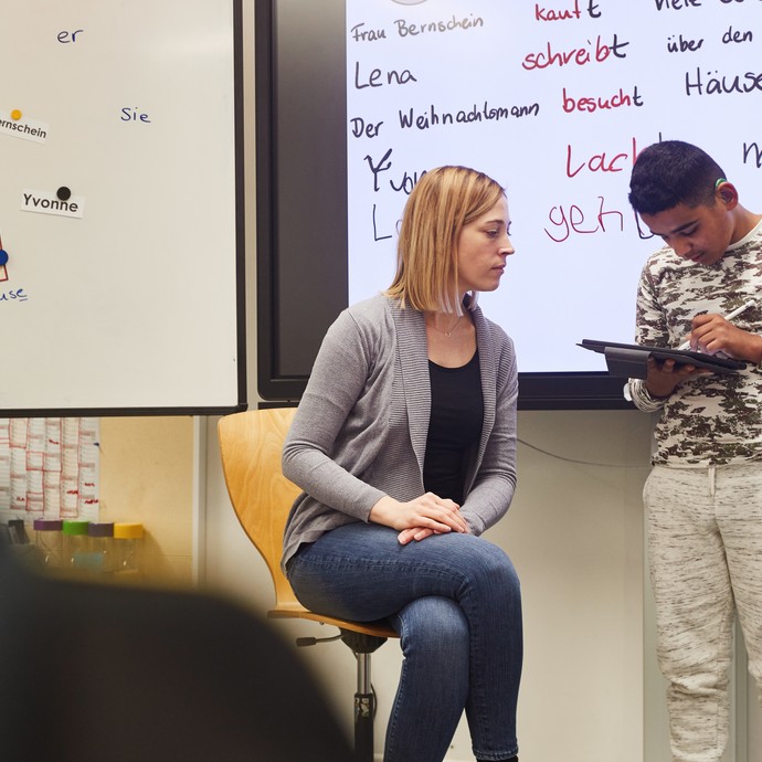 Ein Schüler steht vorne an der Tafel mit einem Tablet, die Lehrerin unterstützt ihn und schaut mit auf das Tablet. (vergrößerte Bildansicht wird geöffnet)