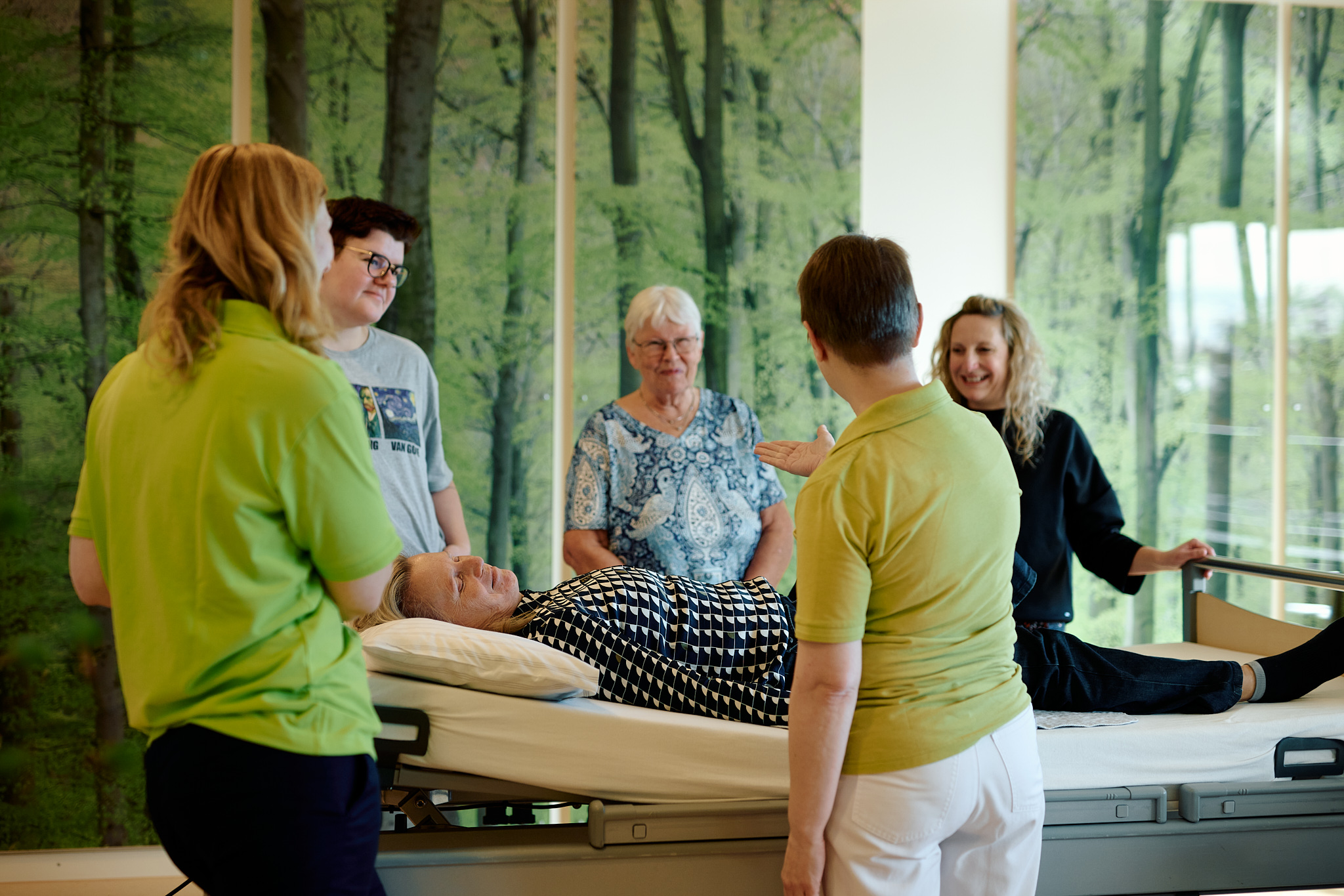 Fünf Menschen stehen im Kreis um eine Pflegebett. Auf dem Bett liegt eine Frau.
