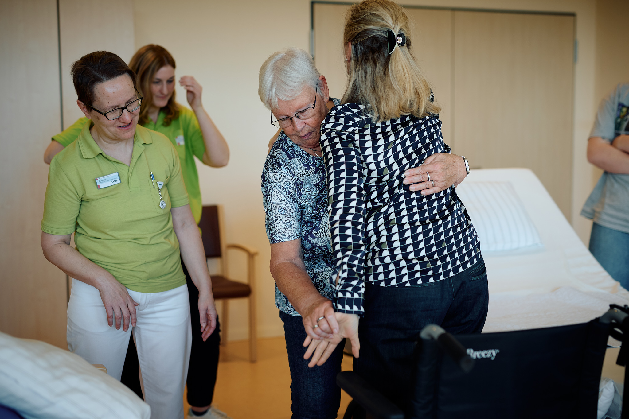 Eine Frau hebt eine andere Frau unter Anleitung eine Pflegekraft in einen Rollstuhl.