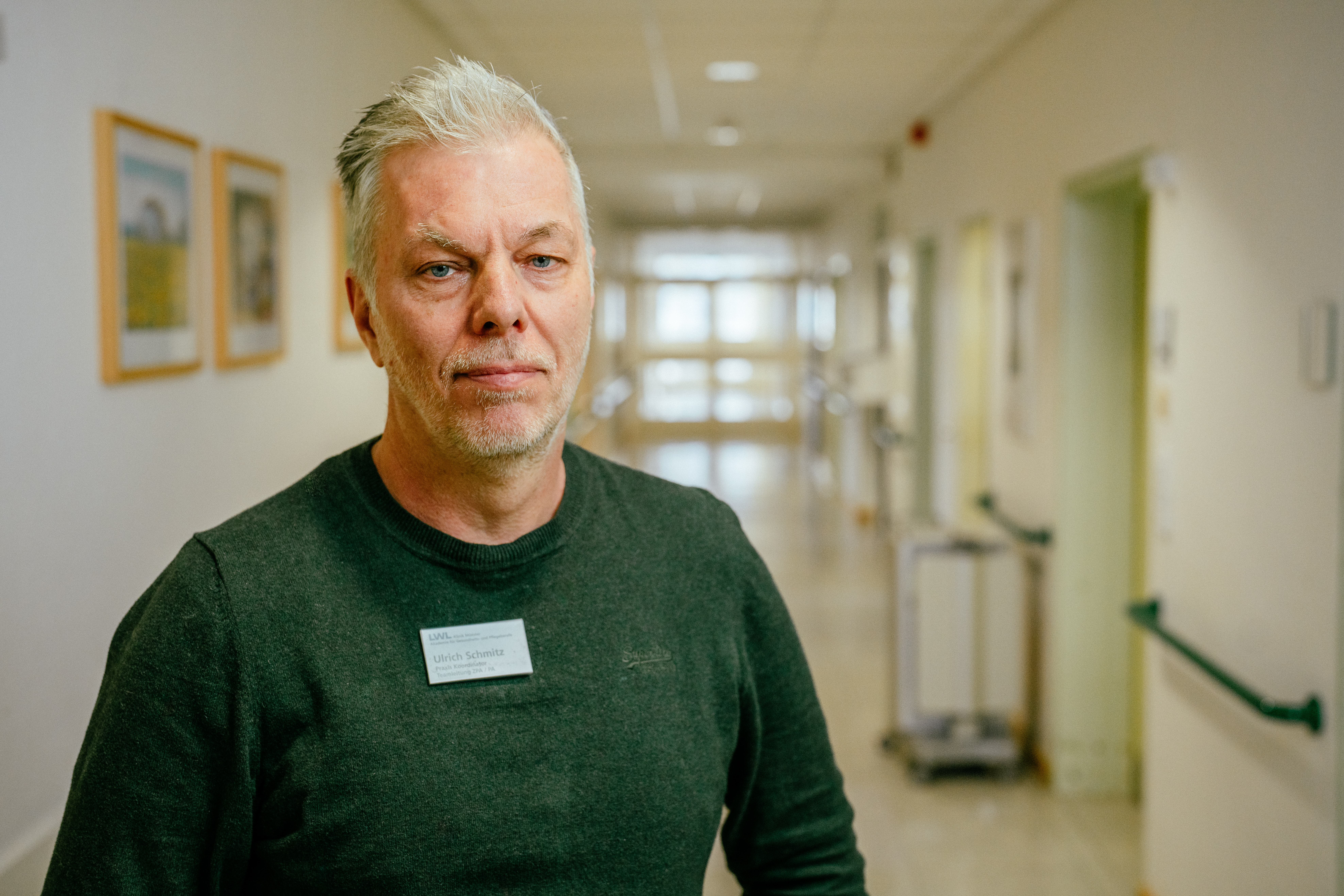 Porträt eines Mannes in grünen Pulli. Er steht auf einem Krankenhausflur.