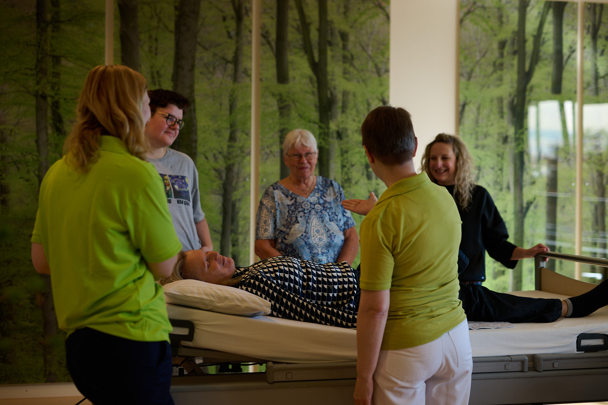 Fünf Menschen stehen im Kreis um eine Pflegebett. Auf dem Bett liegt eine Frau.