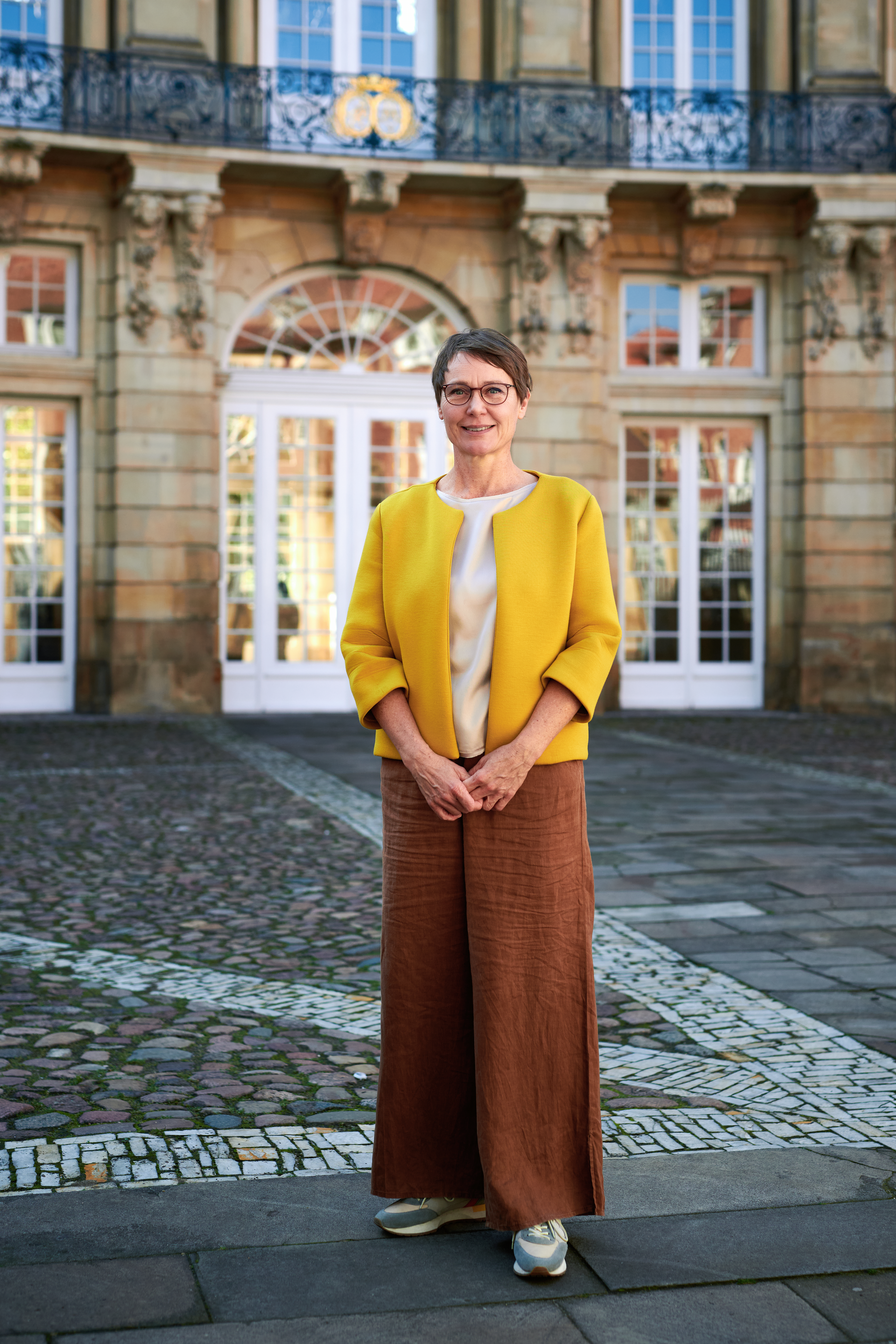 Birte Graue, Frau mit Kurzhaarfrisur und Brille in senffarbenem Oberteil steht vor Erbdrostenhof mir Sandsteinfassade
