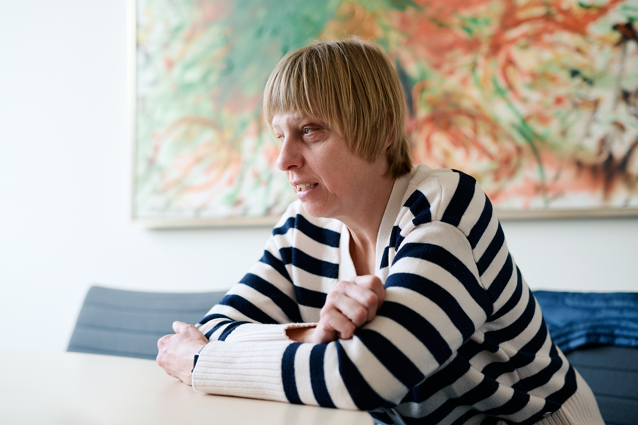 Sabrina Führer im blau-weiß gestreiften Pullover sitzt mit verschränkten Armen an einem Tisch vor einem bunten Gemälde.