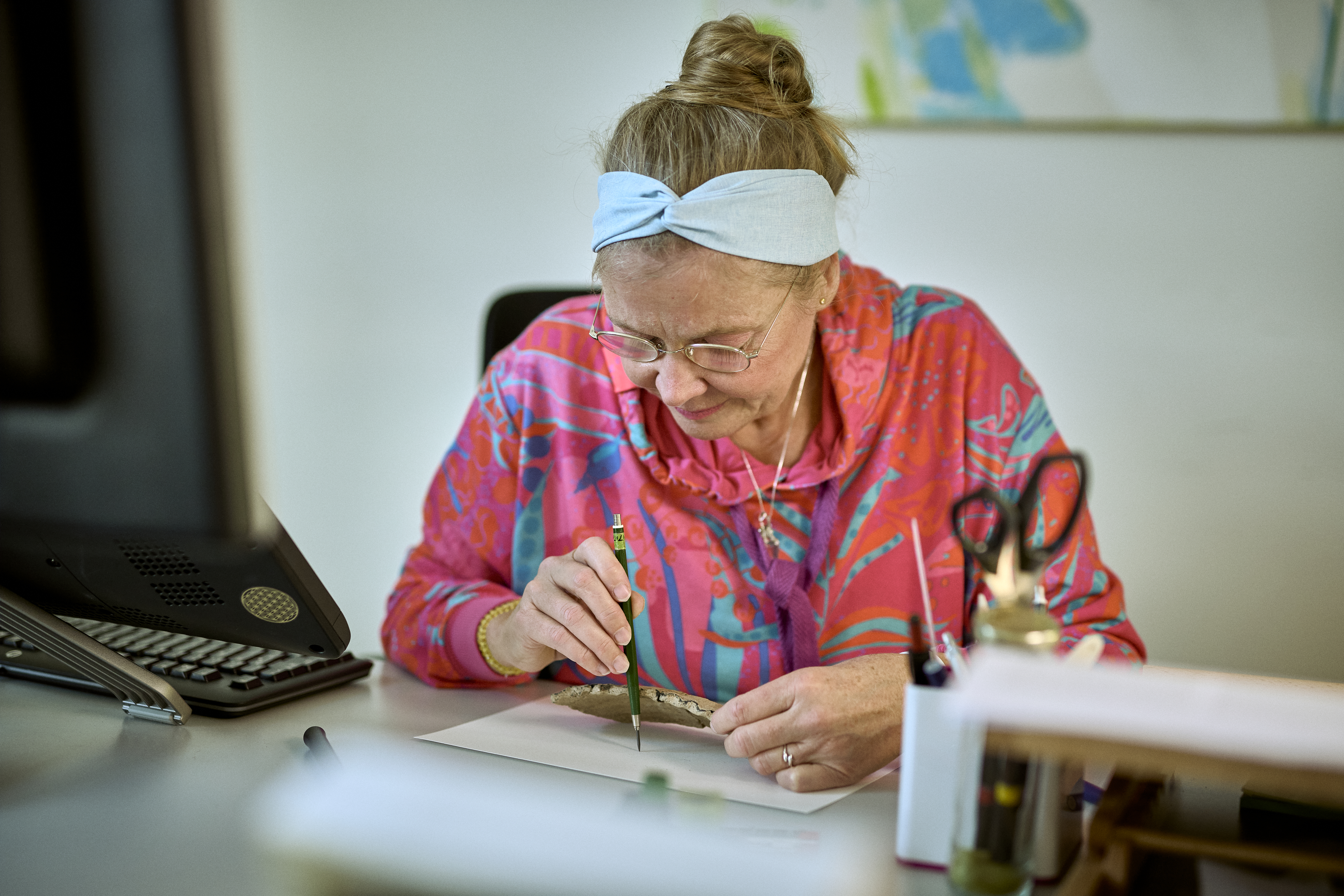 Frau mit korallfarbener Jacke schaut konzentriert auf eine Scherbe in ihrer Hand und malt diese mit einem Stift ab.