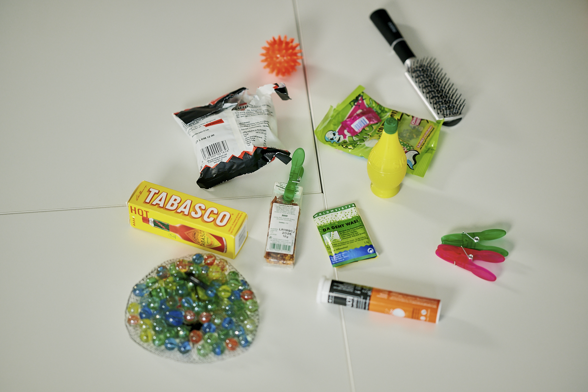 Auf einem Tisch liegen verstreut Gegenstände, u. a. eine Bürste, Wäscheklammern, ein Beutel mit Murmeln und eine Plastikflasche mit Zitronensaft