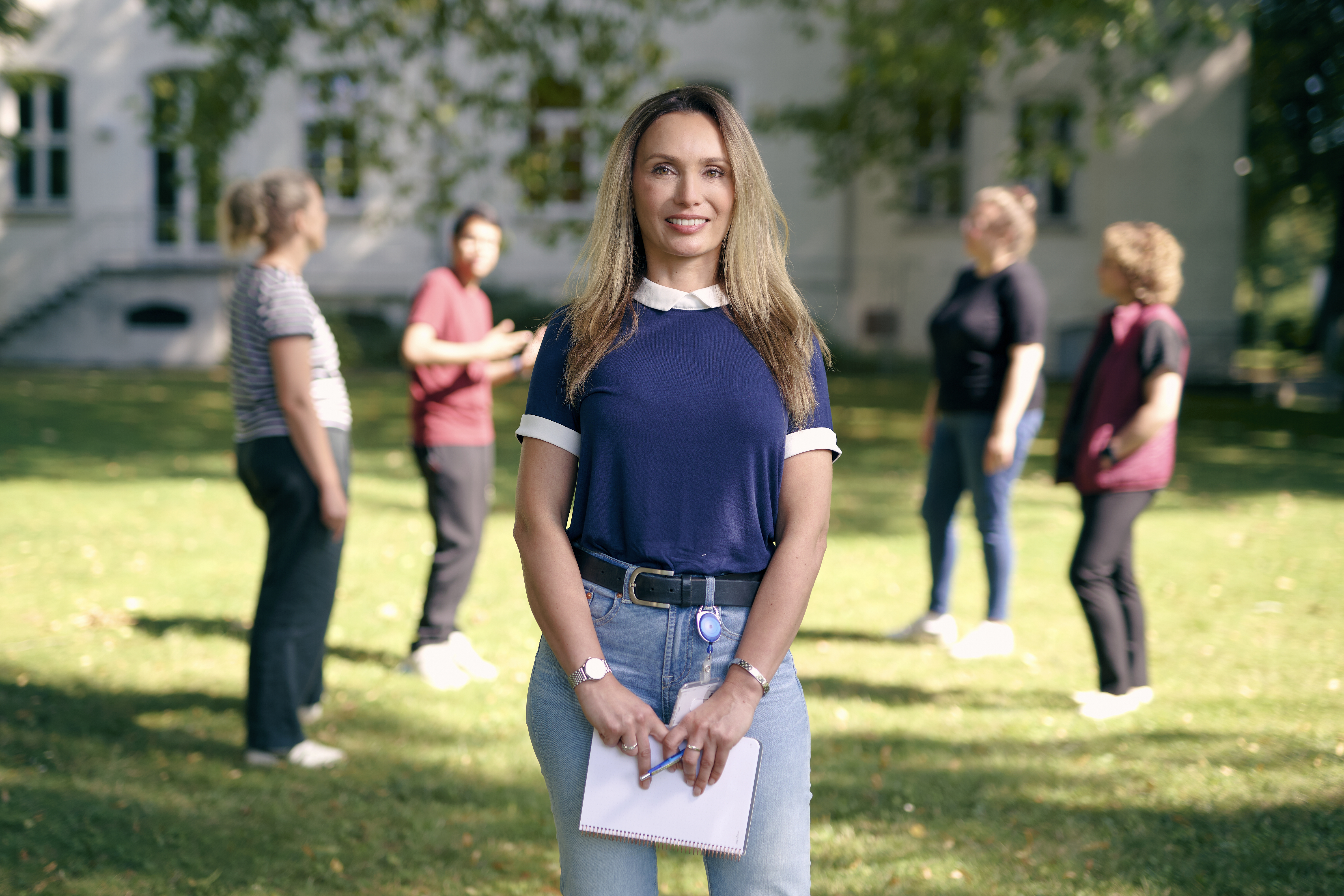 Luljeta Nokaj steht auf einer Wiese vor einem Gebäude. Im Hintergrund sieht man eine Gruppe Menschen, die sich miteinander unterhalten.