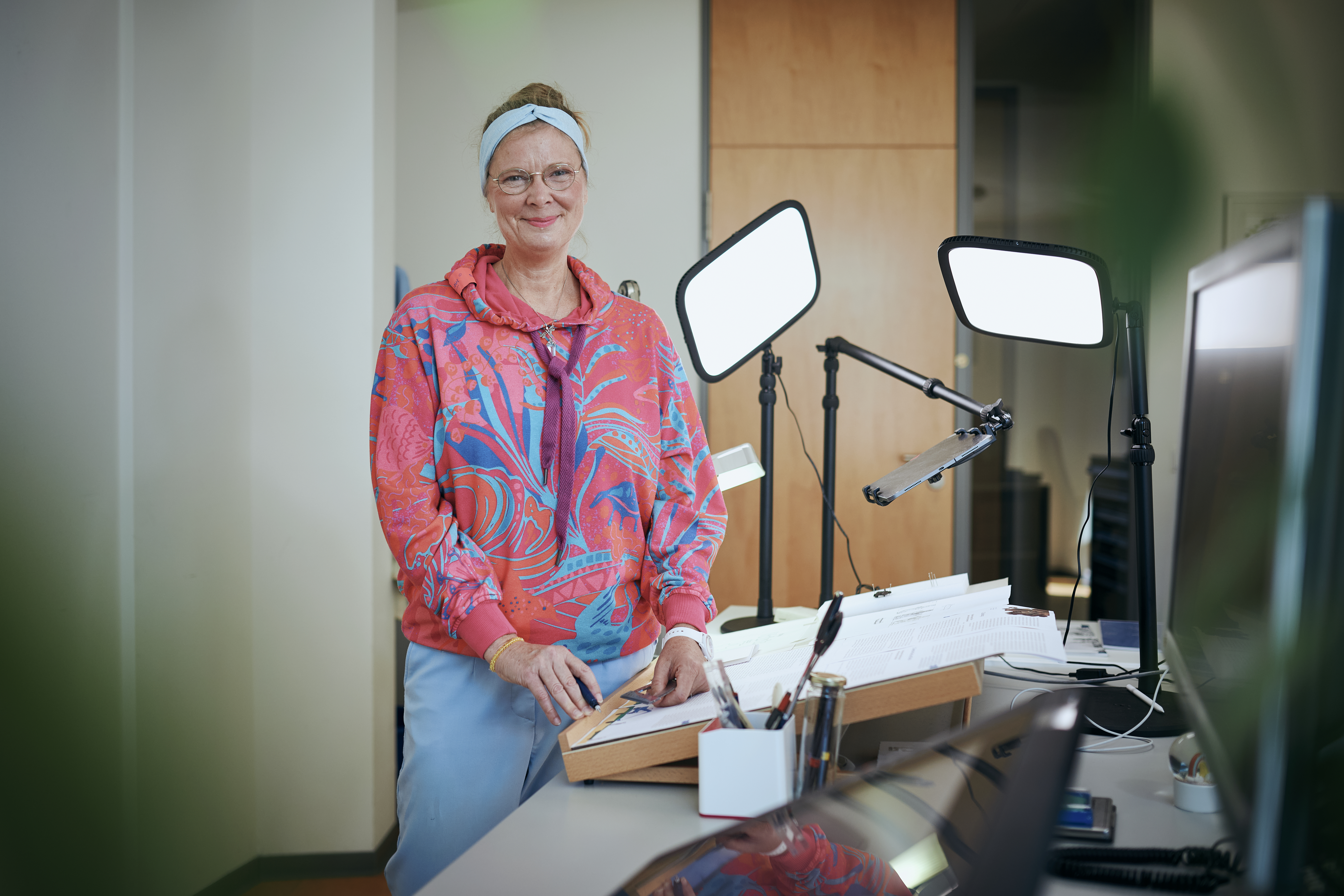 Maike Kloss in korallfarbener Jacke steht neben Zeichentisch mit zwei Arbeitsleuchten und lächelt in die Kamera