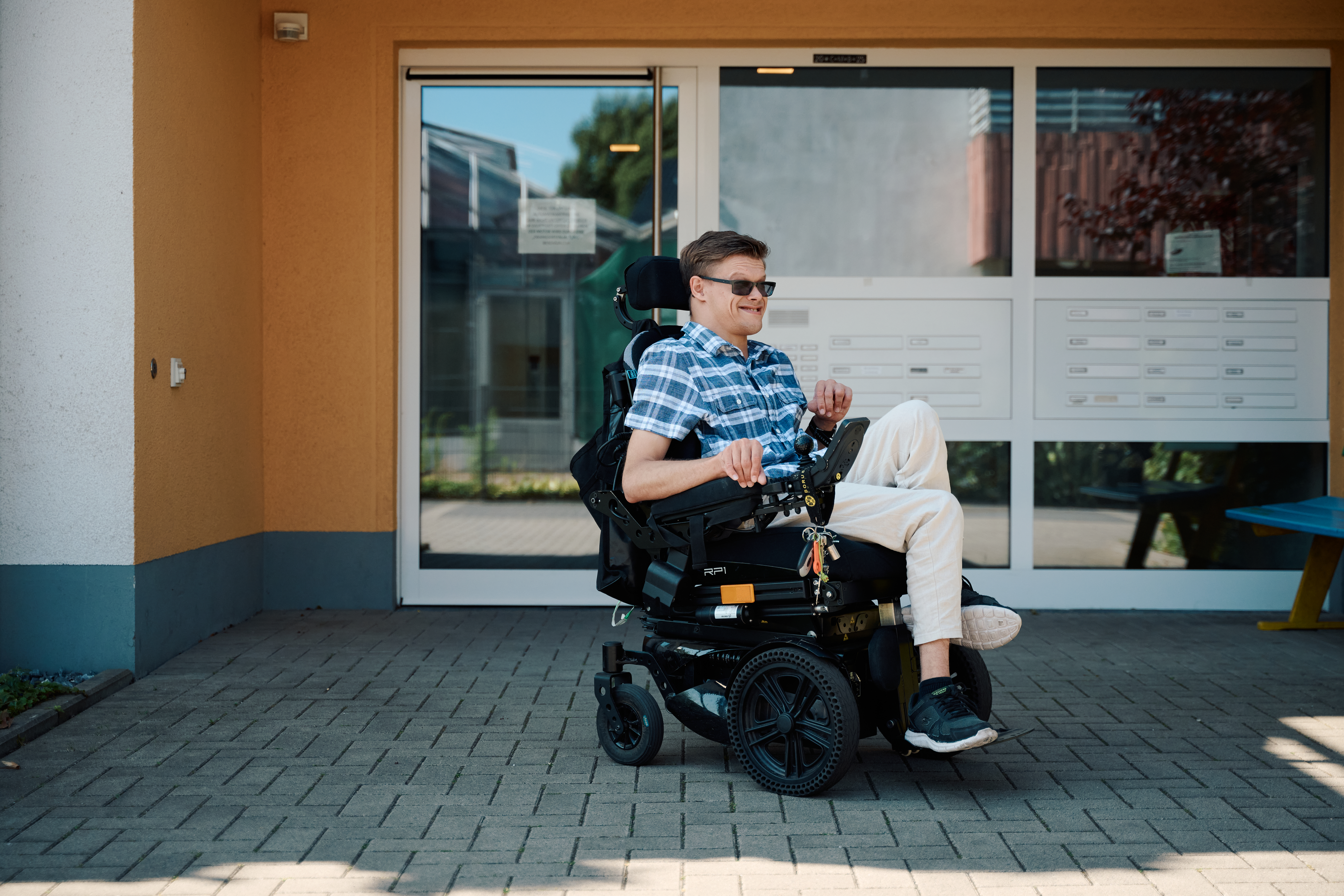 Ein junger Mann im sitzt im elektrischen Rollstuhl vor dem Eingangsbereich eines Mehrfamilienhauses.