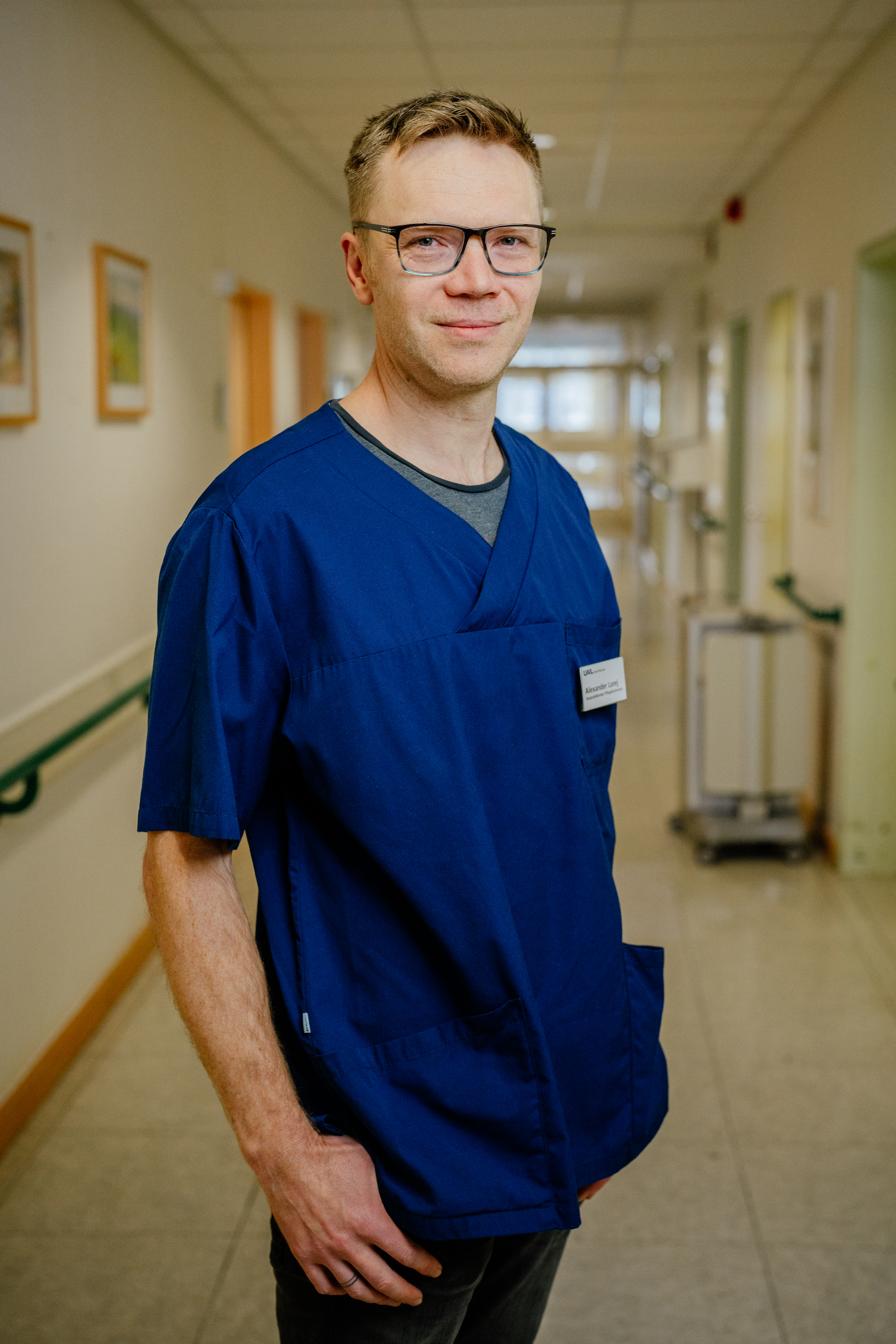 Auszubildender in blauem Pfleger-Oberteil. Er steht auf einem Krankenhausflur.