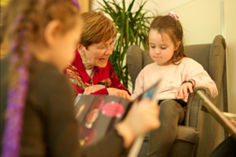 Eine Erzieherin liest gemeinsam mit Kindern. Ein Kind sitzt in einem kleinen Kindersessel.
