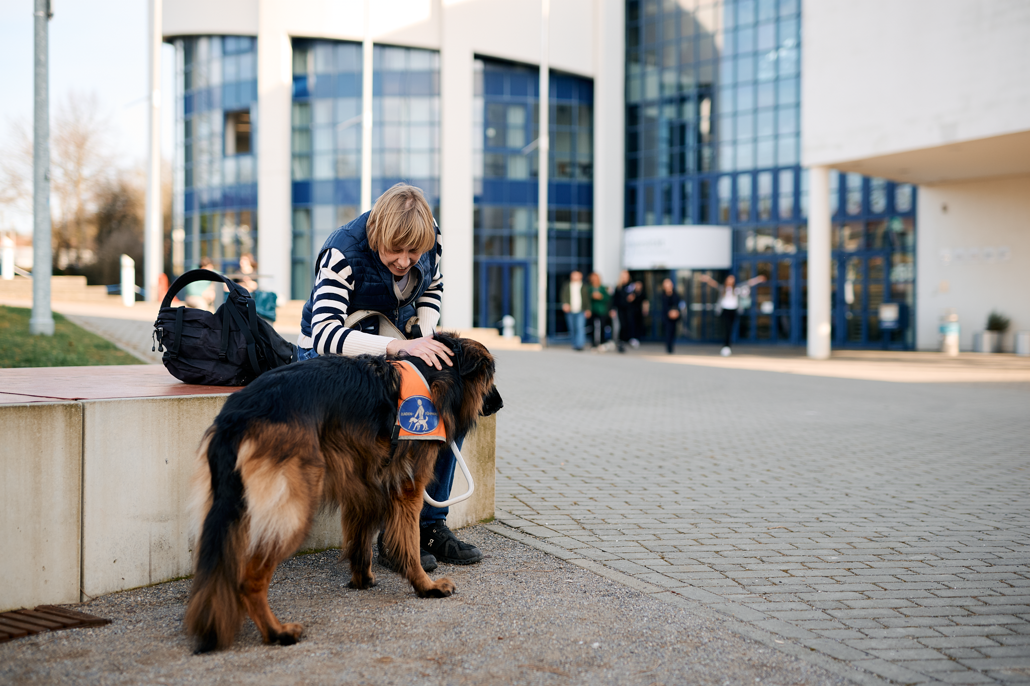 Sabrina Führer sitzt auf einem Betonelement und streichelt Assistenzhünin Reyka. Im Hintergrund kommen Menschen aus dem Hochschulgebäude