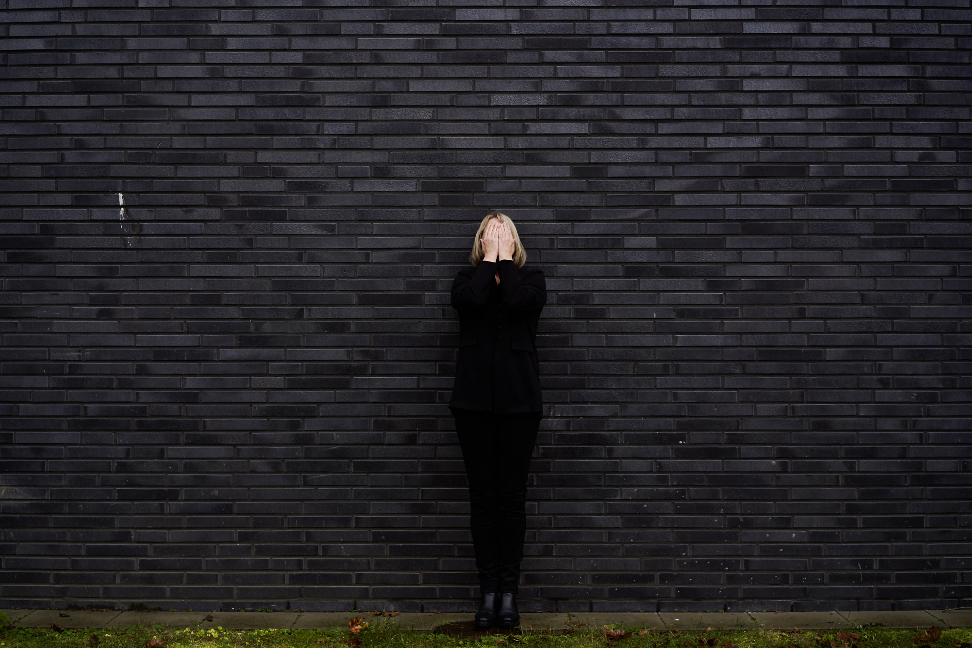 Eine Frau in schwarzer Kleidung steht vor einer schwarzen Wand und hält die Hände vor dem Gesicht.