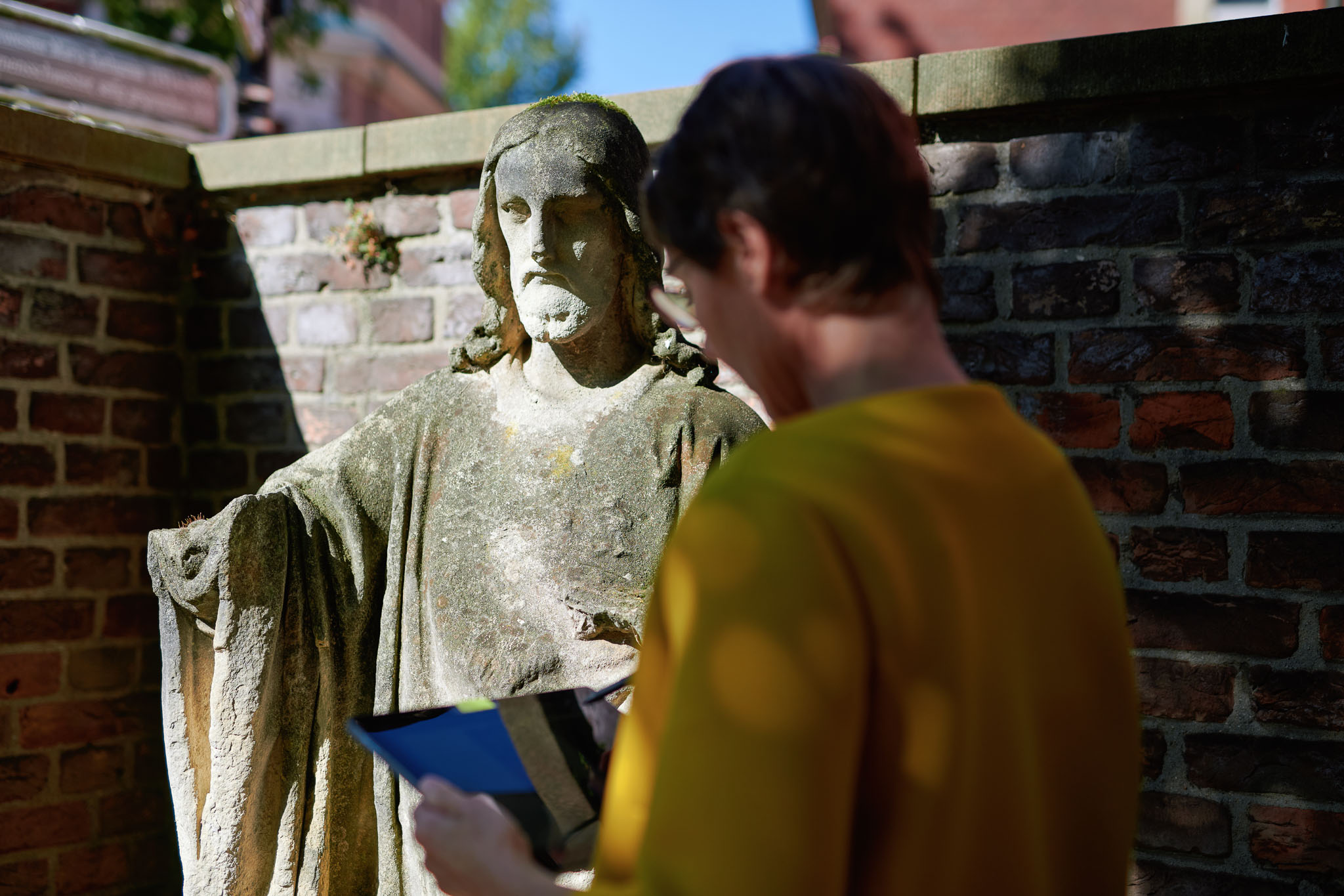 Birte Graue, Frau in senfgelber Jacke betrachtet Tablet, im Hintergrund verwitterte Statue von Mann mit längeren Haaren