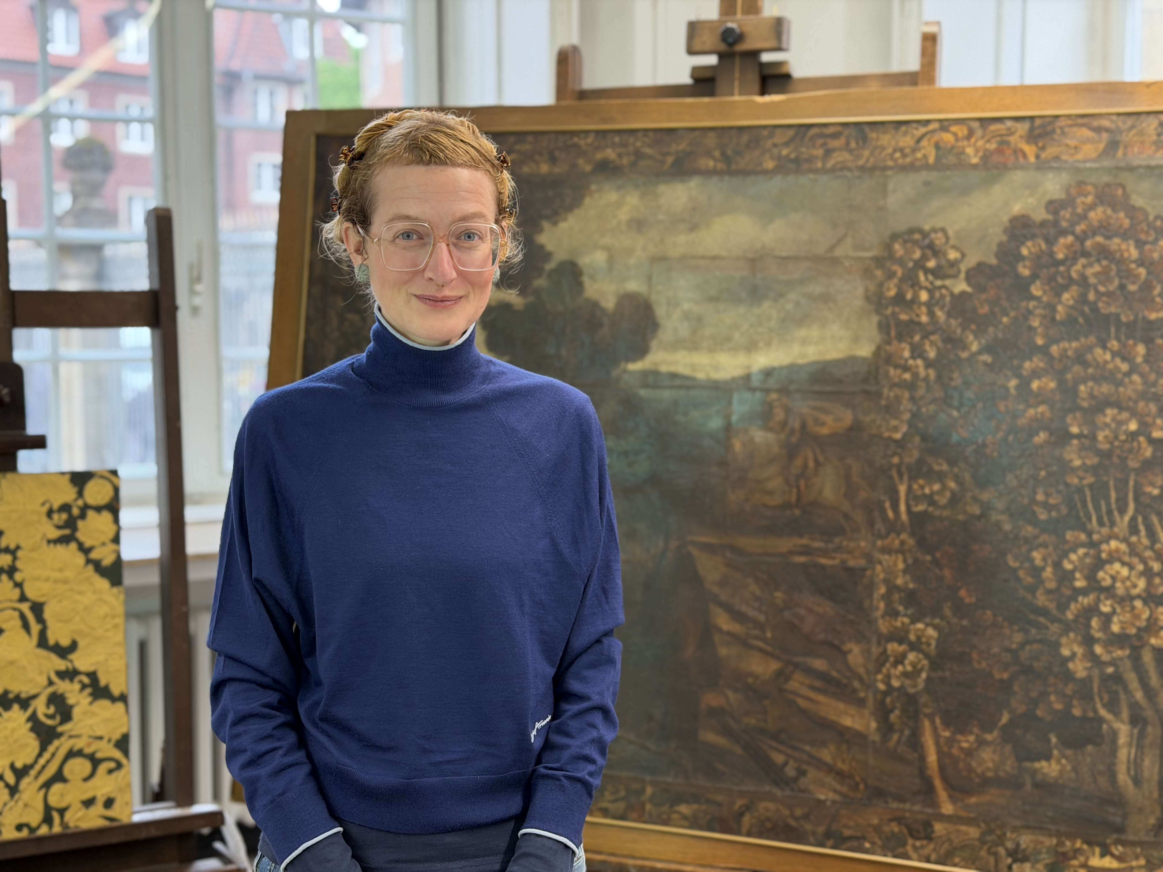 Frau in blauem Pullover steht in einer Restaurationswerkstatt vor einer Staffelei. Man sieht auf dieser einen Teil von einer der Goldledertapeten.