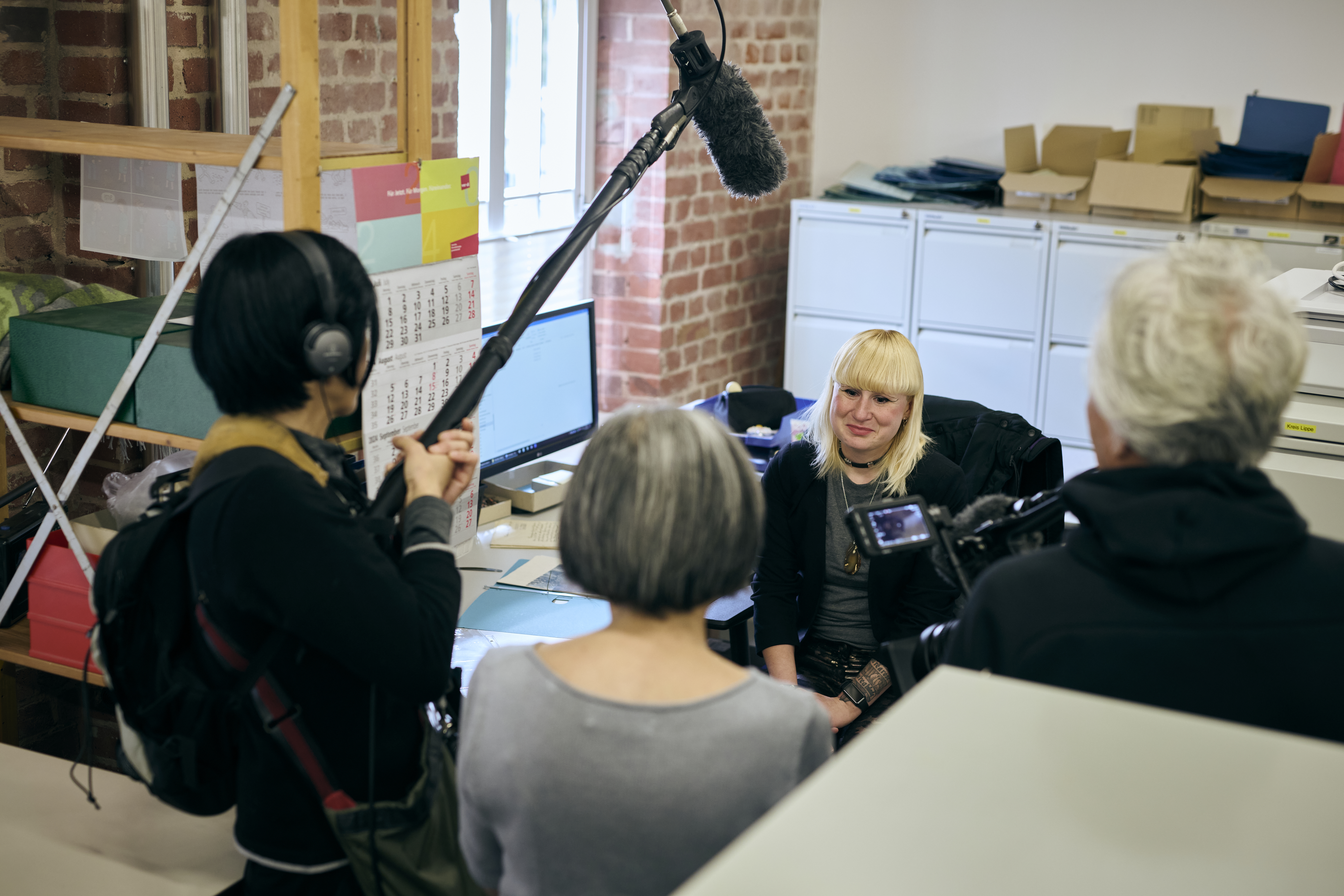 Filmteam filmt eine junge Frau mit blondem Haar an ihrem Schreibtisch am Arbeitsplatz.