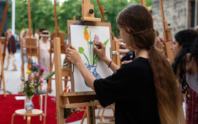 Person mit langen Haaren zeichnet eine Blumenvase auf einer Staffelei im Freien, umgeben von weiteren Personen an Staffeleien.