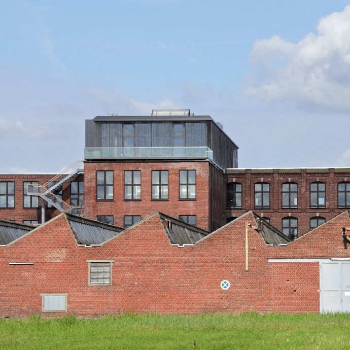Ein altes Industriegebäude aus rotem Backstein mit vielen Fenstern und Dachterasse. Im Vordergrund ist eine Wiese zu sehen (vergrößerte Bildansicht wird geöffnet)