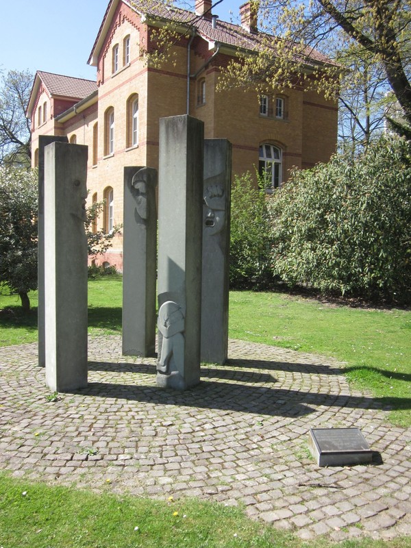 Mahnmal aus mehreren Stelen und Gedenktafel, im Hintergrund Sträucher und eine Haus