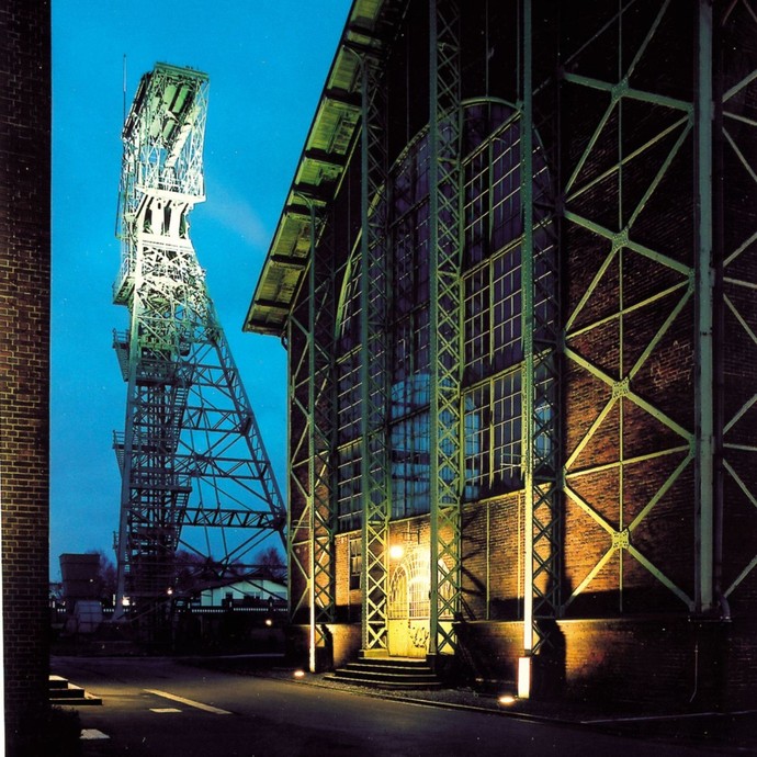 Ein altes Industriegebäude bei Nacht, angestrahlt von Lichtern. Im Hintergrund steht das Fördergerüst der Zeche Zollern (vergrößerte Bildansicht wird geöffnet)