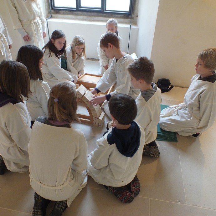 Einige Kinder sitzen in weißen Kutten im Kreis. In der Mitte steht ein Gebile aus Holz mit verschiedenen Bausteinen. (öffnet vergrößerte Bildansicht)