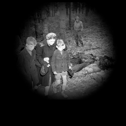 junge Frau führt zwei Kinder an einer Reihe von Leichen vorbei, die auf dem Waldboden liegen, im Hintergrund steht ein amerikanischer Soldat