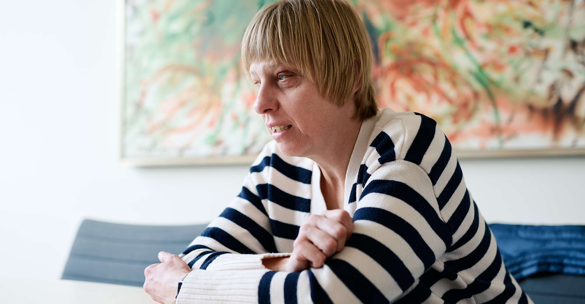 Sabrina Führer im blau-weiß gestreiften Pullover sitzt mit verschränkten Armen an einem Tisch vor einem bunten Gemälde.