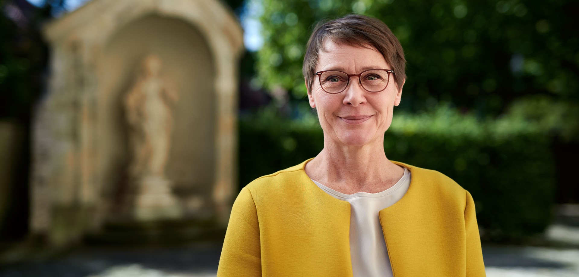 Birte Graue, Frau mit Kurzhaarfrisur und Brille in senffarbenem Oberteil lächelt in die Kamera. Im Hintergrund verschwommen eine eingehauste Sandsteinstatue.
