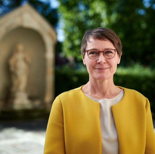 Birte Graue, Frau mit Kurzhaarfrisur und Brille in senffarbenem Oberteil lächelt in die Kamera. Im Hintergrund verschwommen eine Sandsteinstatue. Birte Graue, Frau mit Kurzhaarfrisur und Brille in senffarbenem Oberteil lächelt in die Kamera. Im Hintergrund verschwommen eine Sandsteinstatue.