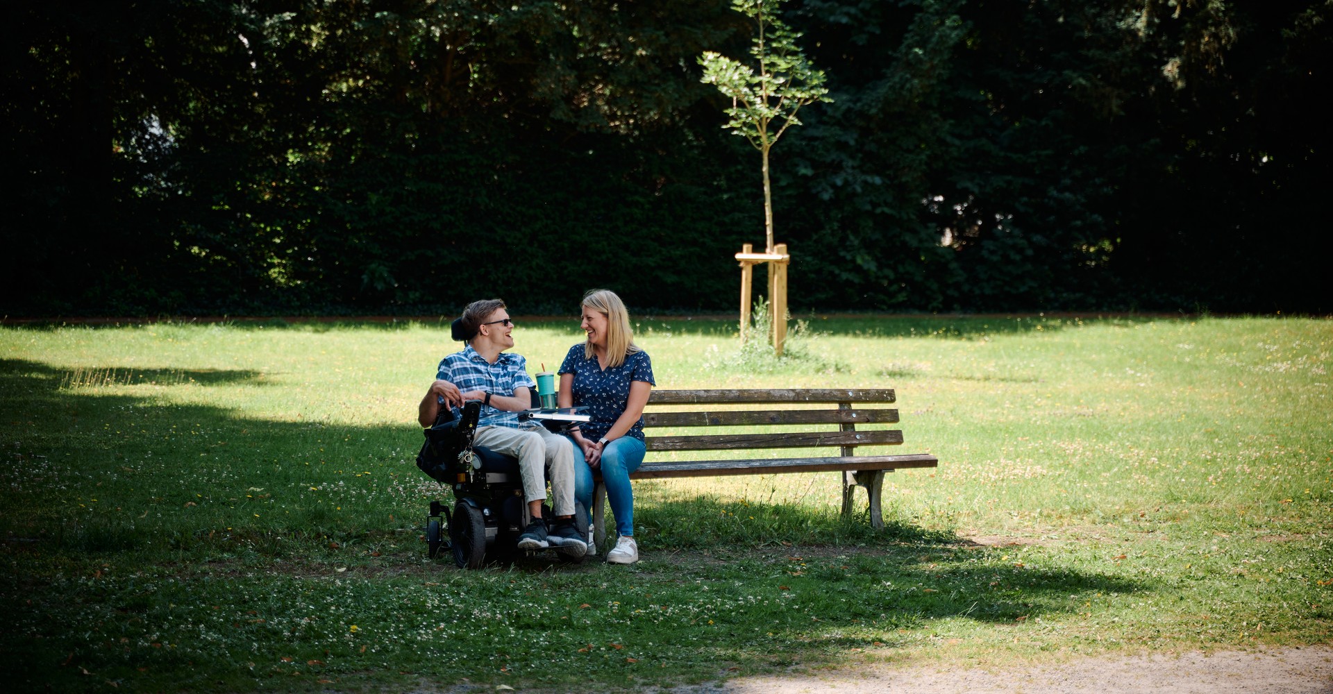 Auf einer Parkbank sitzt eine junge Frau und unterhält sich fröhlich mit einem jungen Mann, der im Rollstuhl direkt neben der Parkbank sitzt.