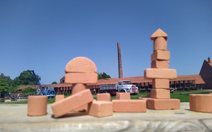 Verschiedene Bauklötze aus Ton stehen auf einem Holzbalken, im Hintergrund sind ein alter Schornstein, ein weißes Fahrzeug und Ziegeldächer zu sehen.
