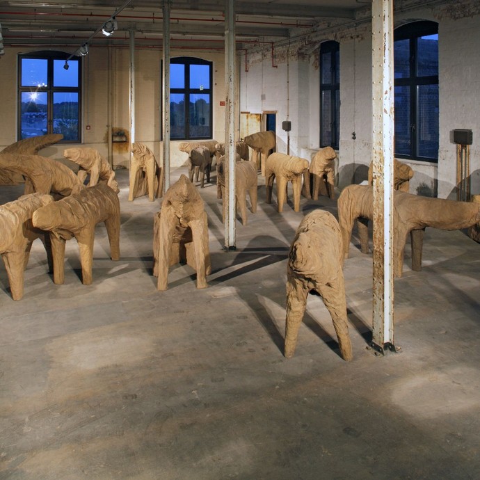 Ein kahler Raum, in welchem viele vierbeinige, aus Holz geschnitzte Wesen aufgestellt sind (vergrößerte Bildansicht wird geöffnet)