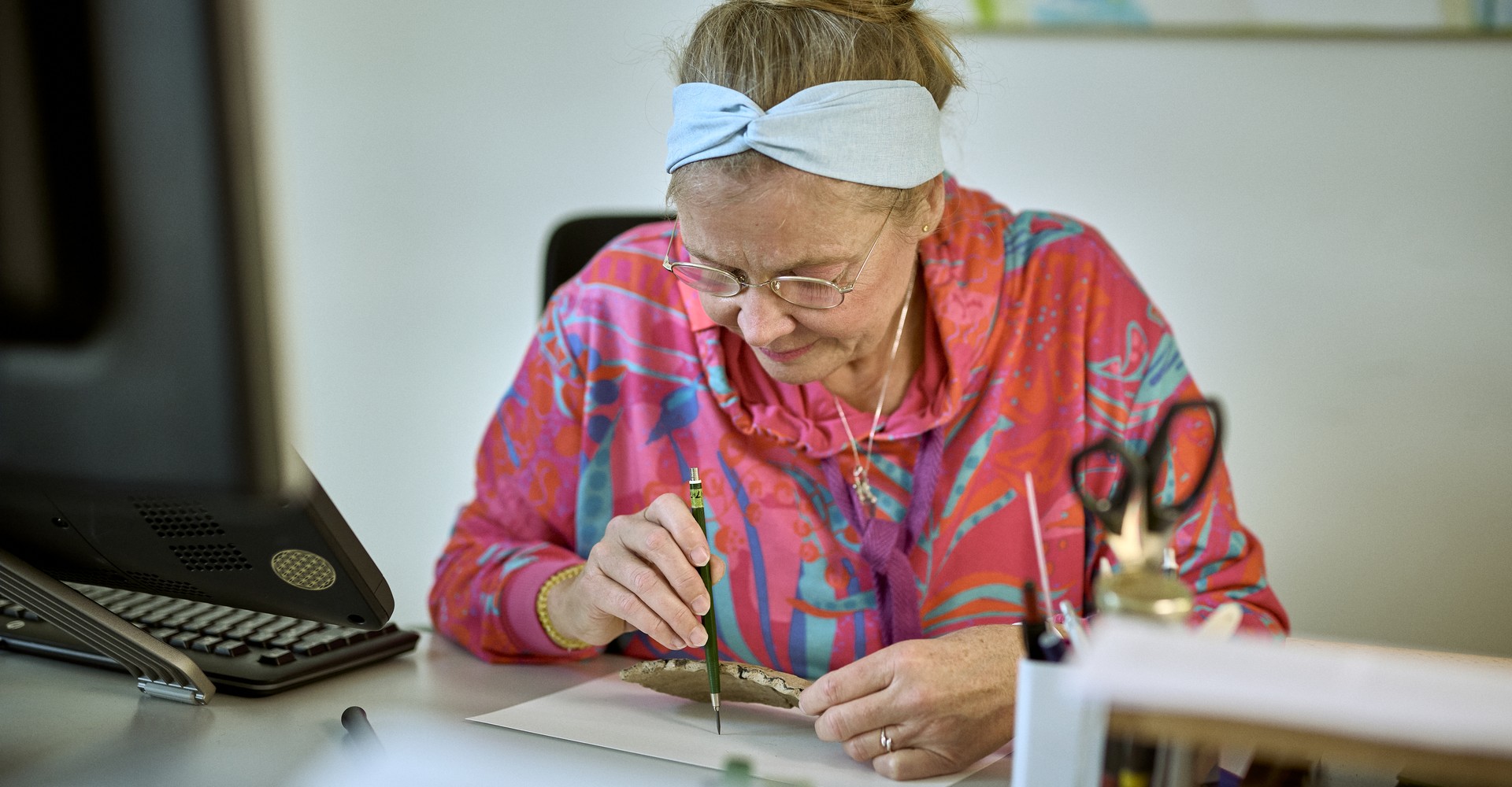 Frau mit korallfarbener Jacke schaut konzentriert auf eine Scherbe in ihrer Hand und malt diese mit einem Stift ab.