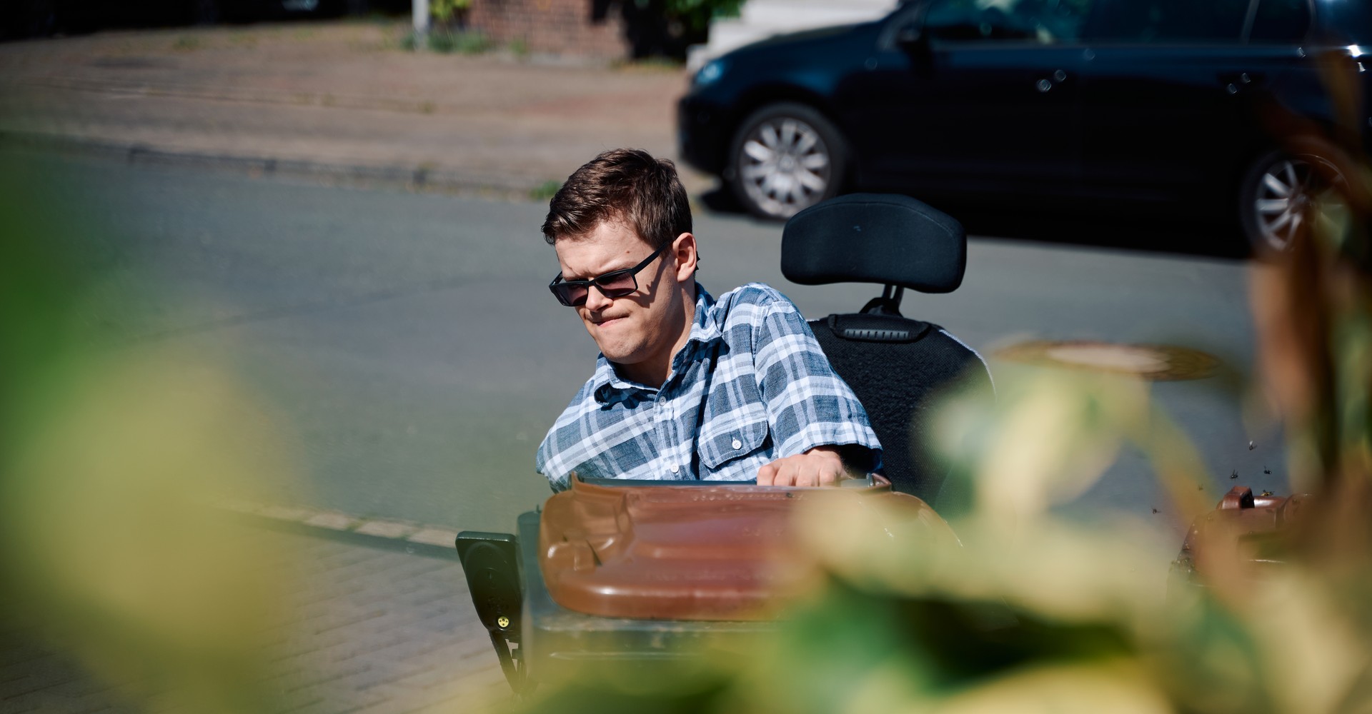 Ein junger Mann mit Sonnenbrille im Rollstuhl zieht eine Bio-Mülltonne.