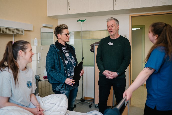 Die Ausbilder geben der Auszubildenden eine Rückmeldung, alle stehen neben einem Krankenhausbett, in dem die Patientin liegt.