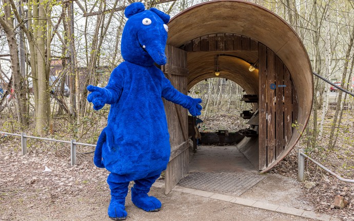 Person in blauem Tierkostüm einer Ratte mit großen weißen Augen steht vor einem halbrunden, hölzernen Tunnelzugang.