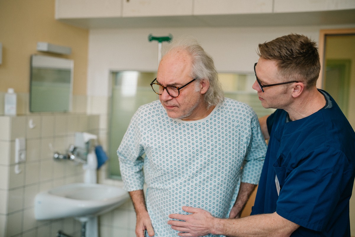 Ein Pfleger in blauem Krankenhaus-Oberteil stützt einen älteren Mann im Patientenkittel. Ein Pfleger in blauem Krankenhaus-Oberteil stützt einen älteren Mann im Patientenkittel.