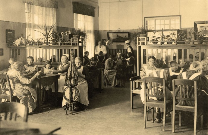 Foto aus alten Zeiten: Frauen sitzen an Tischen und arbeiten, zum Beispiel am Spinnrad