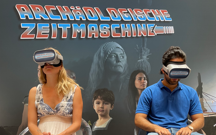 Zwei Personen mit VR-Brillen sitzen auf Stühlen vor einem Plakat mit dem Schriftzug 'ARCHÄOLOGISCHE ZEITMASCHINE'. Die Personen tragen VR-Brillen.