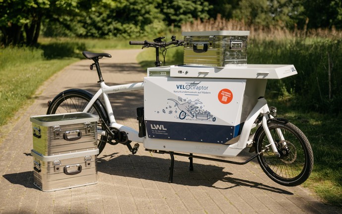 Lastenfahrrad mit großem weißen Kasten und mehreren silbernen Transportboxen auf einem Weg im Park