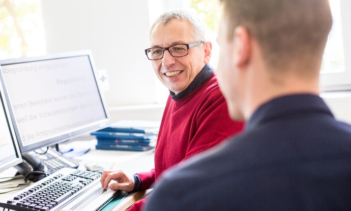 Ein älterer Mann sitzt vor einem Computer mit Braillezeile und bekommt Unterstützung von einem jüngeren Mann Ein älterer Mann sitzt vor einem Computer mit Braillezeile und bekommt Unterstützung von einem jüngeren Mann