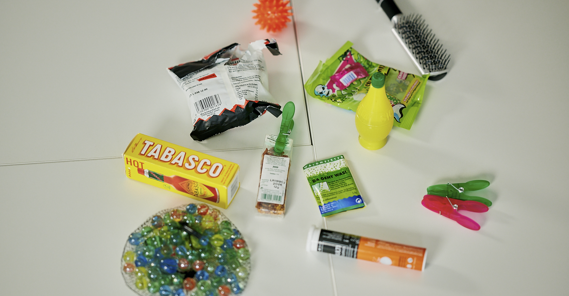 Auf einem Tisch liegen verstreut Gegenstände, u. a. eine Bürste, Wäscheklammern, ein Beutel mit Murmeln und eine Plastikflasche mit Zitronensaft