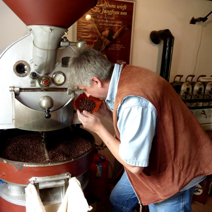 Ein Röster prüft den Röstgrad der Kaffeebohnen. Im Hintergrund steht die Maschine, welche die Bohnen röstet. (vergrößerte Bildansicht wird geöffnet)