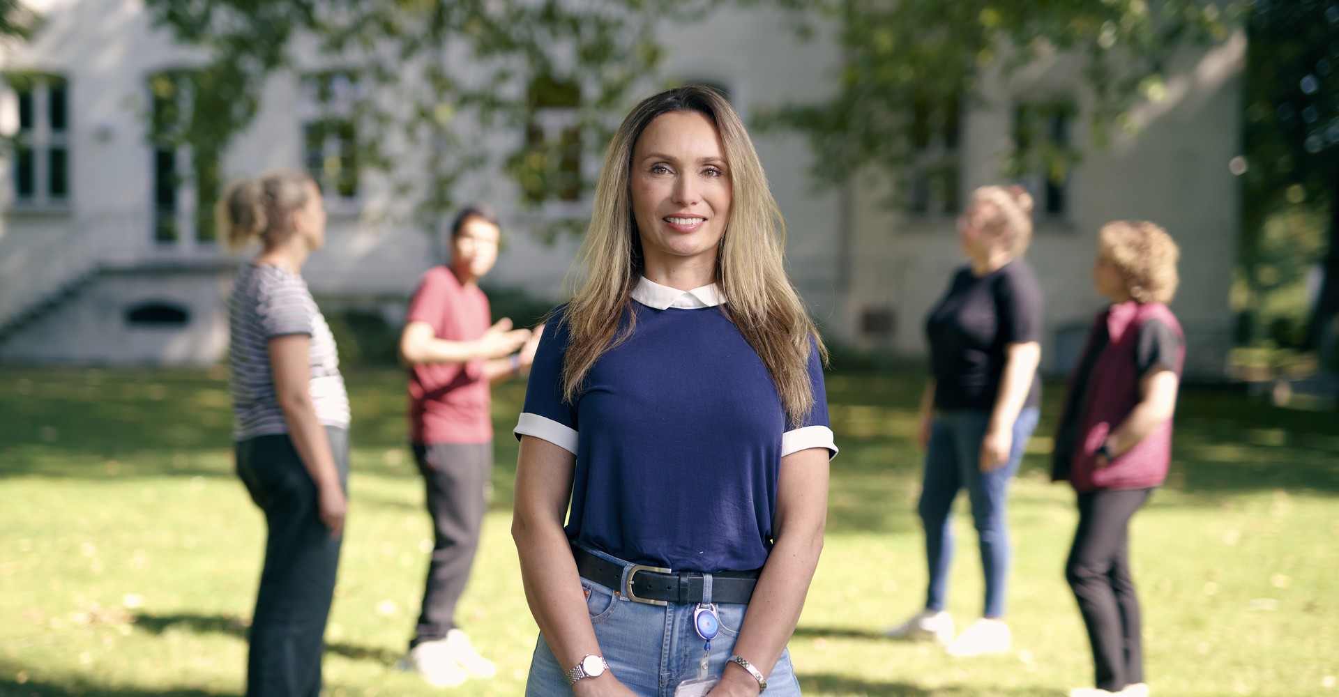Luljeta Nokaj steht auf einer Wiese vor einem Gebäude. Im Hintergrund sieht man eine Gruppe Menschen, die sich miteinander unterhalten.