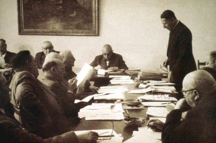 Mehrere Männer sitzen mit Dokumenten an Tischen