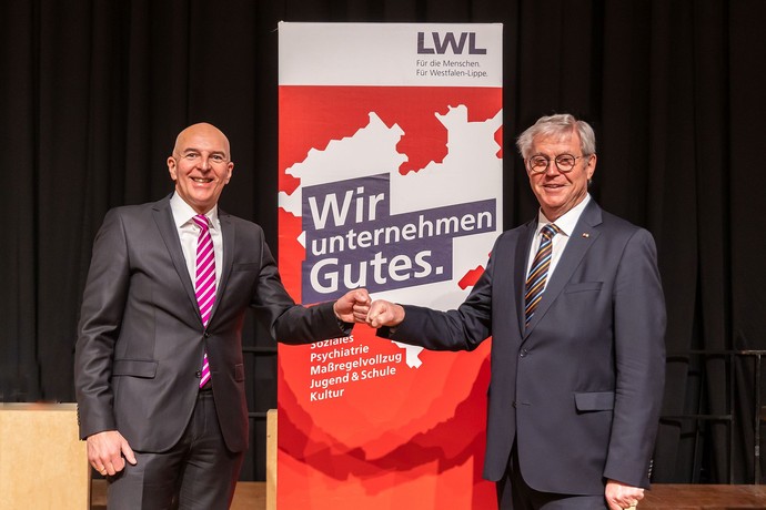 Klaus Baumann gratuliert Dr. Georg Lunemann vor einem Plakat mit der Aufschrift "Wir unternehmen Gutes".