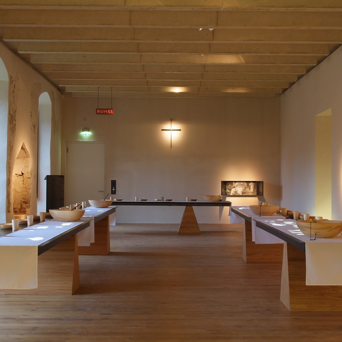 Ein großer Raum mit Steinmauern und drei länglichen Tischen. Auf den Tischen steht Geschirr aus Holz. (vergrößerte Bildansicht wird geöffnet)