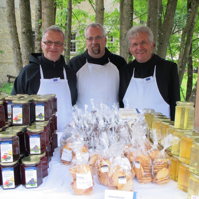 Drei Männer in weißer Schürze stehen hinter einem Tisch, an dem Marmelade, Kekse und Honig verkauft wird. (vergrößerte Bildansicht wird geöffnet)