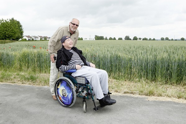 Vater schiebt den Rollstuhl seines Sohnes über einen Feldweg, beide lachen.